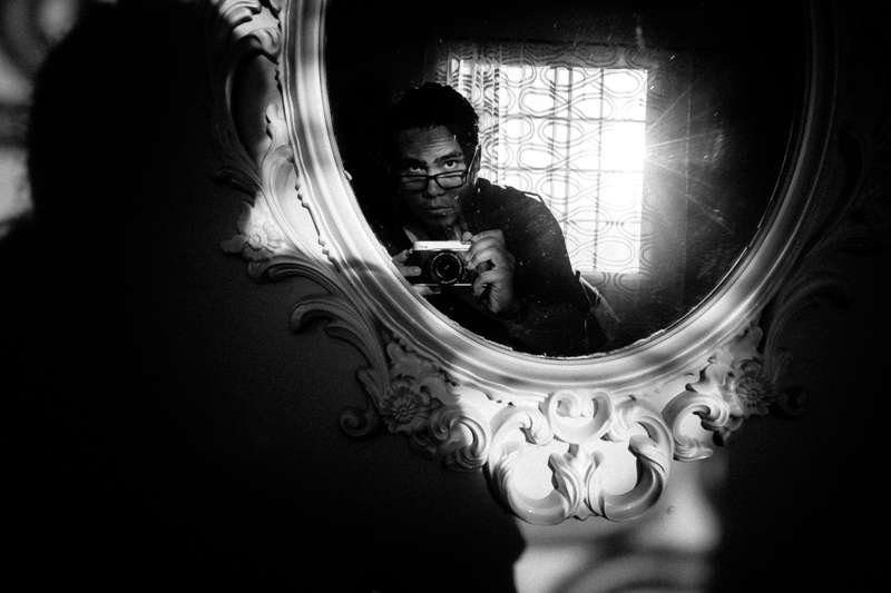 Black and white self-portrait of a photographer holding a small camera, reflected in an ornate round mirror in a dark room