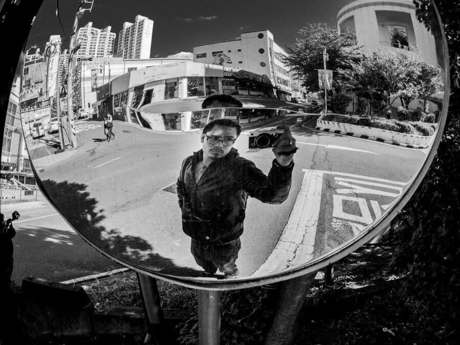 Photographer seen in a round convex traffic mirror on a street corner in Korea, holding a camera up with one hand