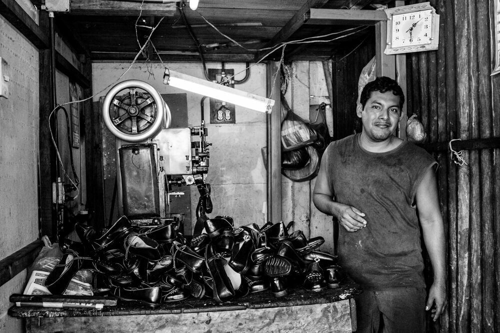 Black and white photo of a man standing beside a pile of shoes in a small shop in Nicaragua, with a clock and a scale on the wall