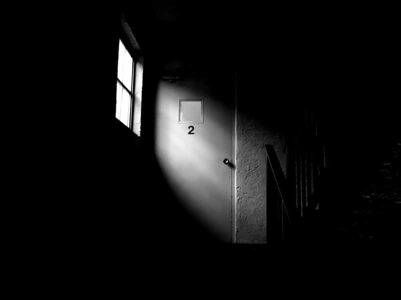Black and white photo of a dark hallway where a single window throws a patch of light on a door marked 2