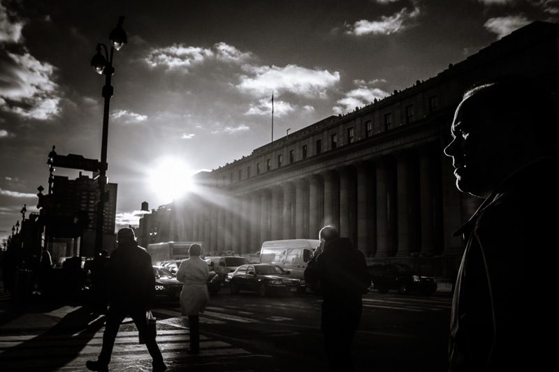 Black and white street photo of people crossing toward a columned building with the sun flaring behind them and a man's profile in the foreground