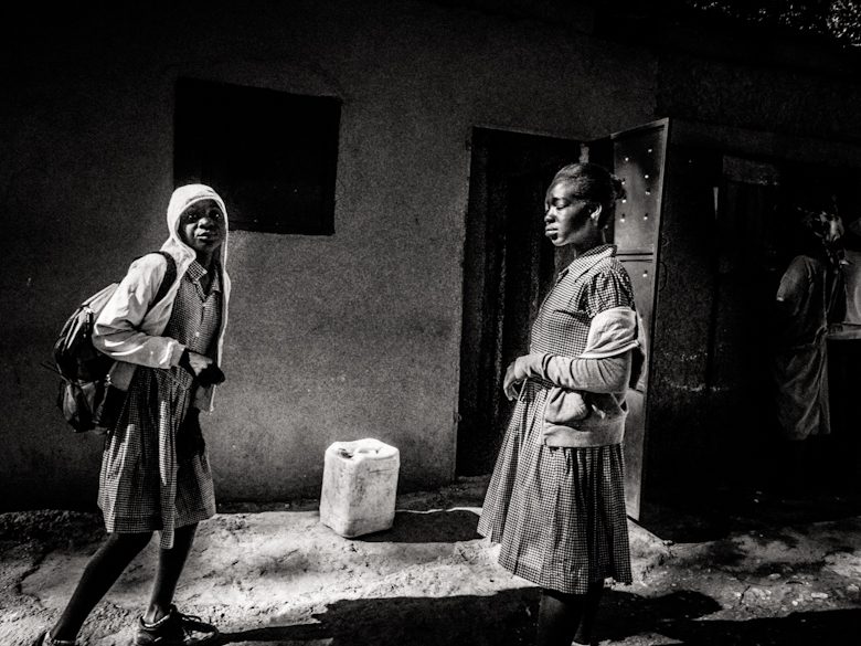 Black and white photo of a schoolgirl walking toward a woman standing in a doorway in Haiti, with a jerrycan between them