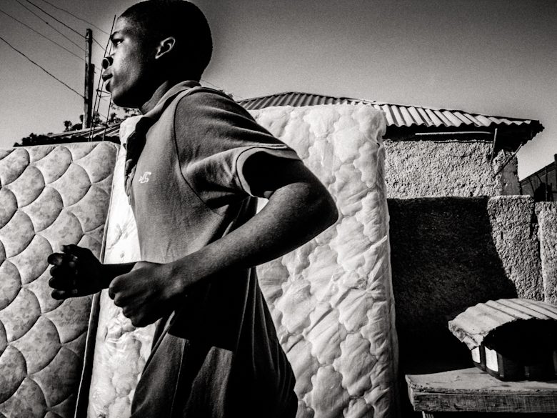 Black and white street photo of a young man running past a mattress propped against a wall in Haiti