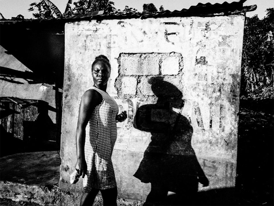 Black and white photo of a woman standing beside a whitewashed wall with a strong shadow of another person on it, Haiti