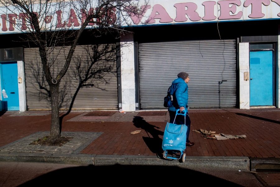 Street photo of a woman in a blue jacket pulling a blue shopping cart past blue shop doors