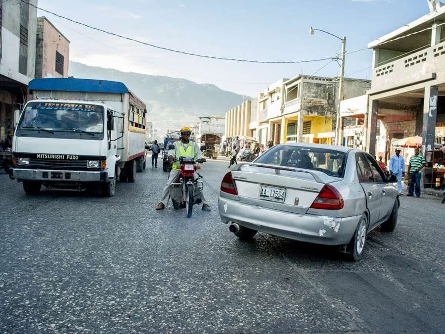 Busy street with a motorbike taxi between a truck and a silver car, colour street photo